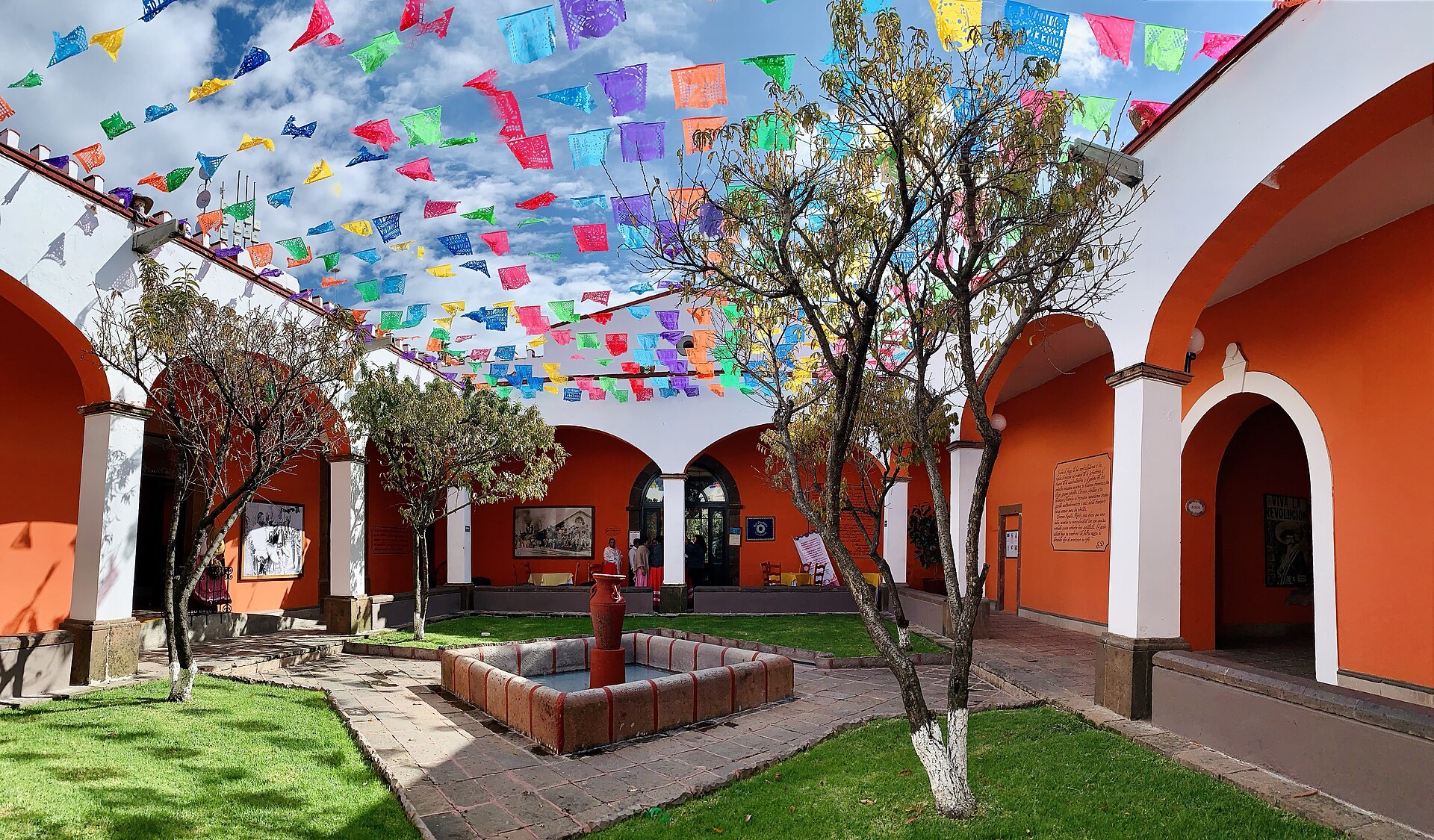 Imagen ilustrativa: patio de hacienda mexicana con arcos naranjas, fuente, jardín y papel picado de colores