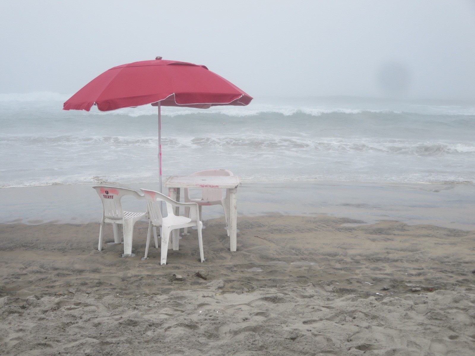 Sombrilla roja con mesa y sillas blancas en la playa de Rosarito en un día nublado