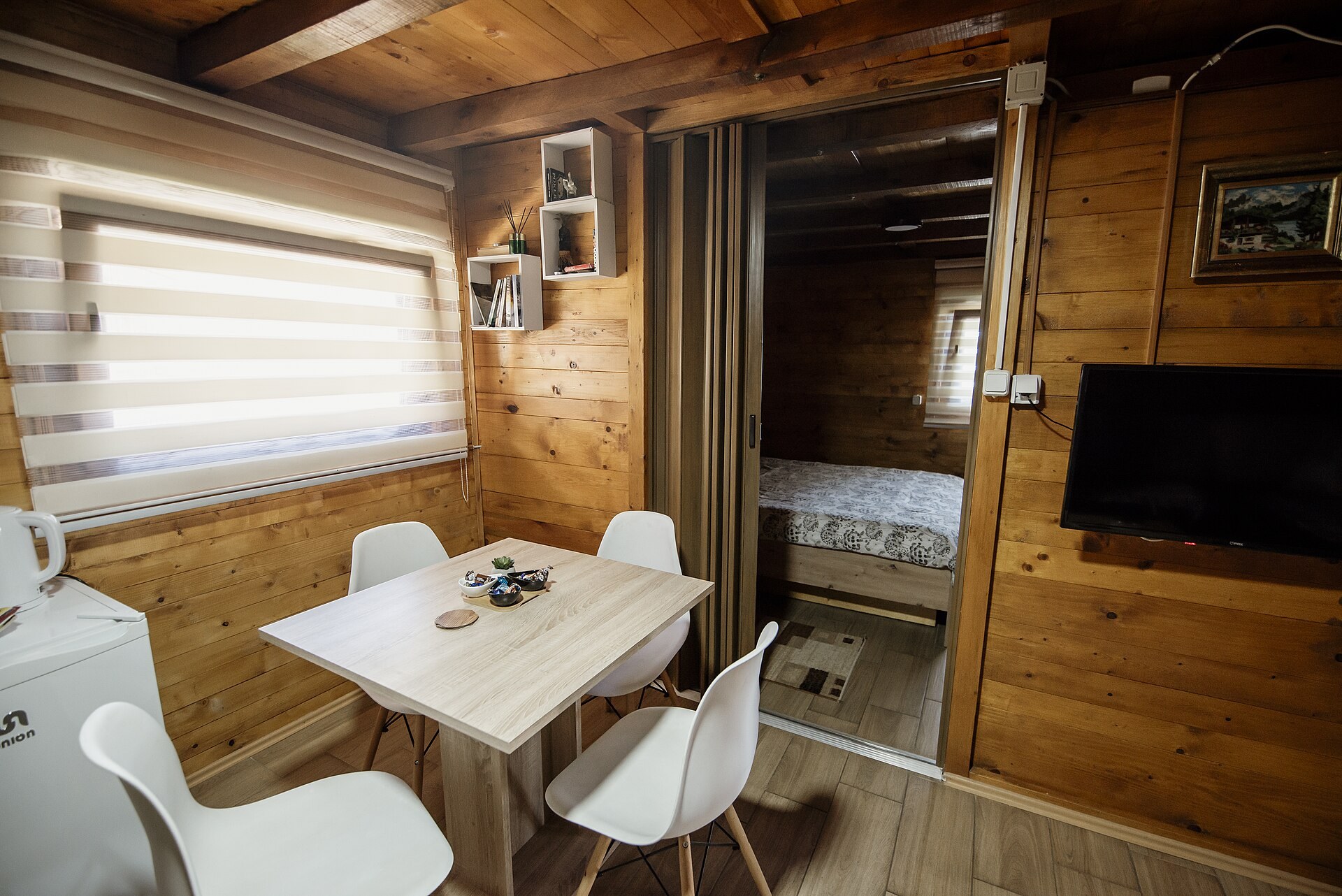 Imagen ilustrativa: interior de cabaña de madera con comedor blanco para cuatro, cocineta y acceso a la recámara